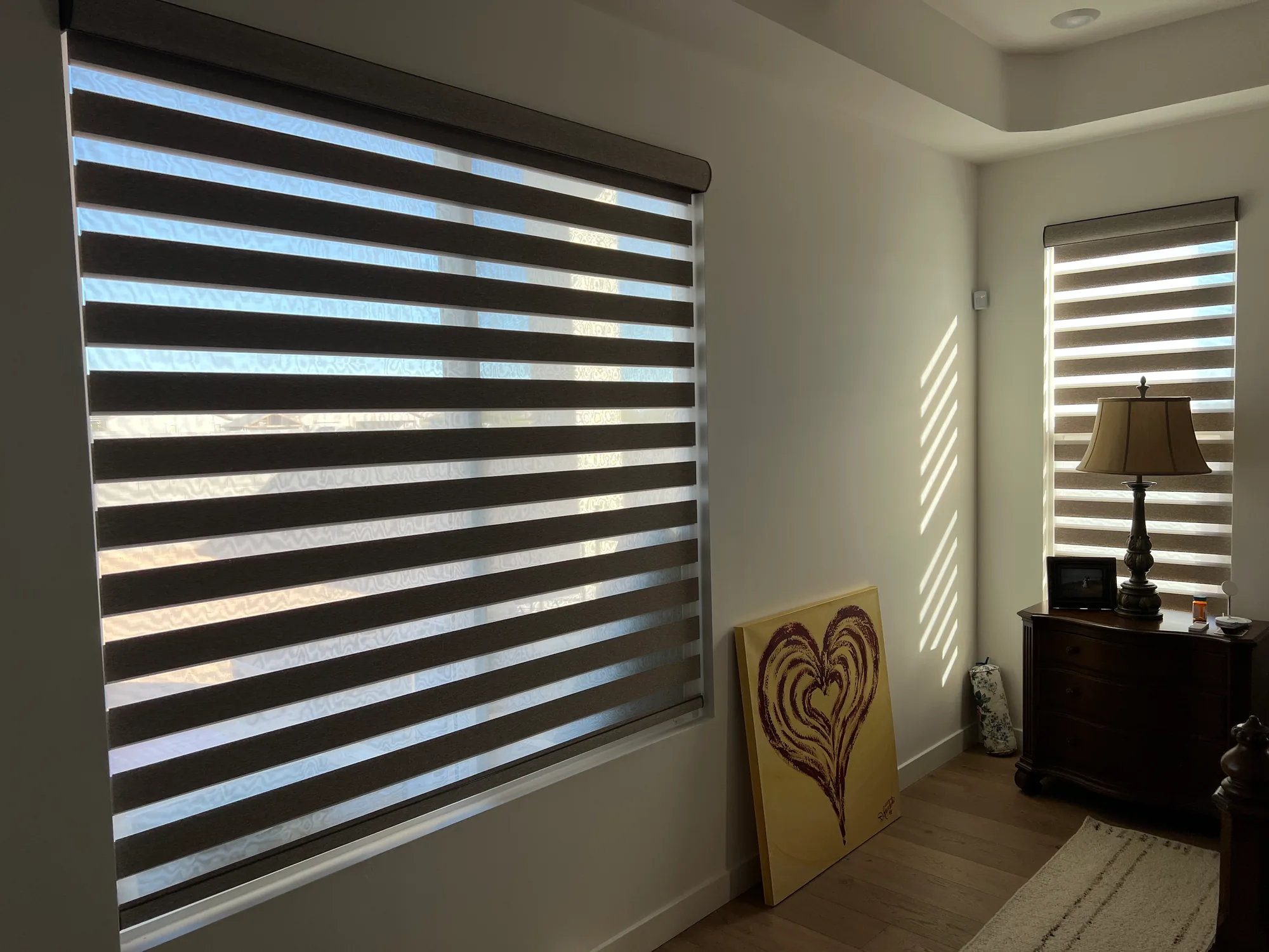 Dark zebra shades on two bedroom corner windows casting striped daylight onto a white wall.