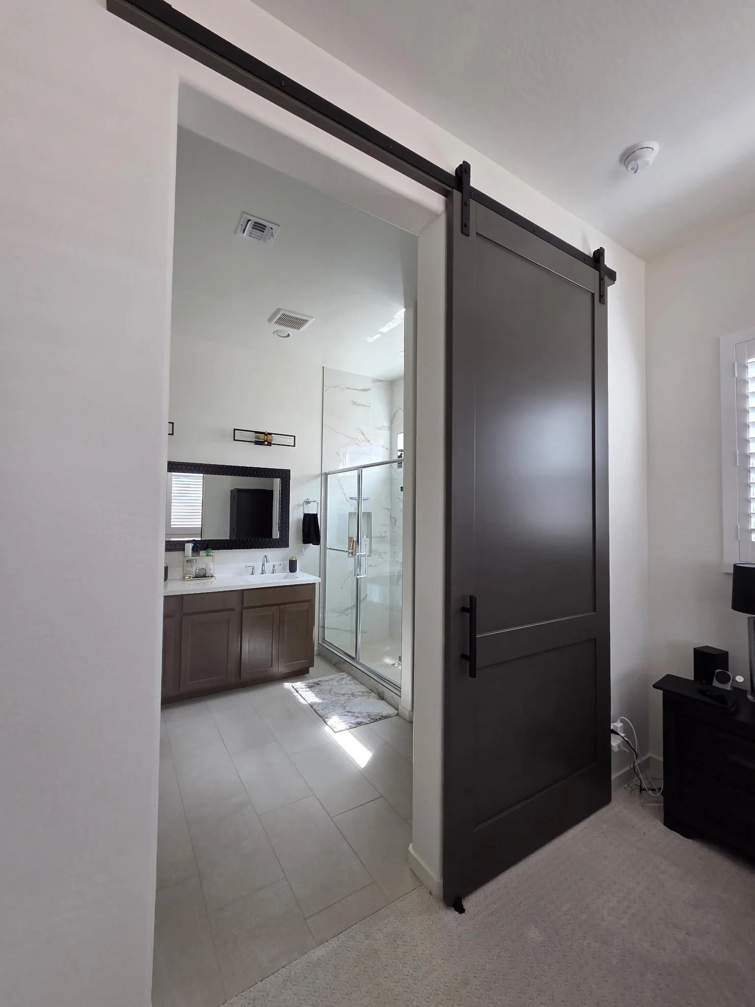 Two Panel Shaker barn door, dark finish with a tall upper panel and shorter lower panel, partly open at a bathroom entrance on an exposed black rail